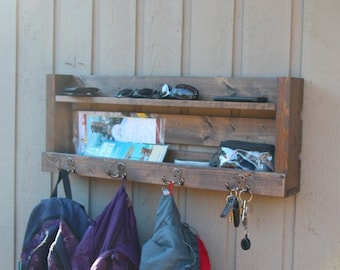 May include: A rustic wooden wall shelf with two shelves and hooks. The shelf is stained a dark brown and has a distressed finish. It has a variety of items on it, including sunglasses, books, a clear plastic bag, and keys. There are three backpacks hanging on the hooks.