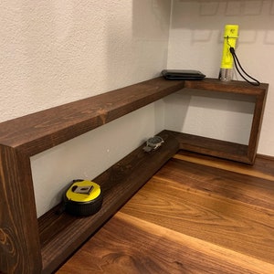 Pu&ograve; includere: Un ripiano d'angolo in legno marrone scuro con due ripiani. Il ripiano superiore ha un portafoglio nero e una torcia elettrica gialla. Il ripiano inferiore ha un orologio e un metro a nastro giallo.