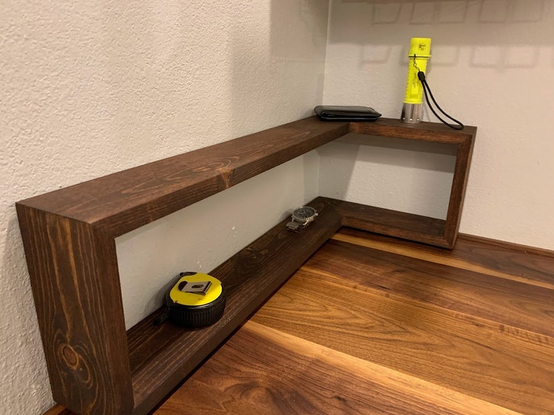 Pu&ograve; includere: Un ripiano d'angolo in legno marrone scuro con due ripiani. Il ripiano superiore ha un portafoglio nero e una torcia elettrica gialla. Il ripiano inferiore ha un orologio e un metro a nastro giallo.