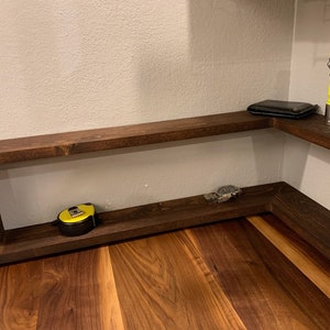 Pu&ograve; includere: Un ripiano d'angolo in legno marrone scuro con due ripiani. Il ripiano superiore ha un portafoglio nero e una torcia elettrica gialla. Il ripiano inferiore ha un metro a nastro giallo e un orologio argentato.