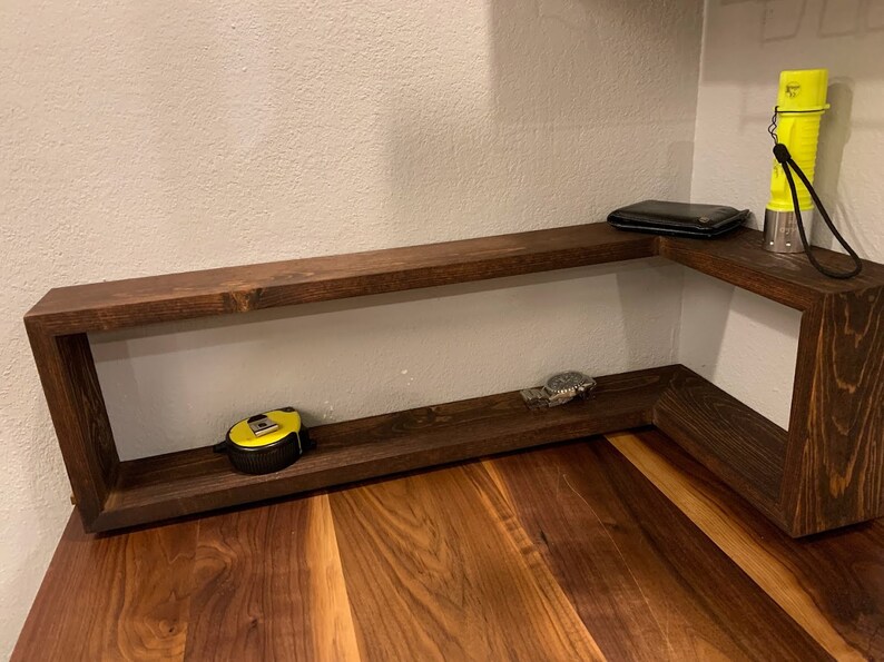 Pu&ograve; includere: Un ripiano d'angolo in legno marrone scuro con due ripiani. Il ripiano superiore ha un portafoglio nero e una torcia elettrica gialla. Il ripiano inferiore ha un metro a nastro giallo e un orologio argentato.