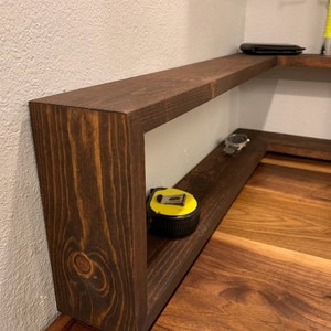 Pu&ograve; includere: Un ripiano d'angolo in legno marrone scuro con due ripiani. Il ripiano superiore ha un portafoglio nero sopra. Il ripiano inferiore ha un metro a nastro giallo e un orologio argentato.