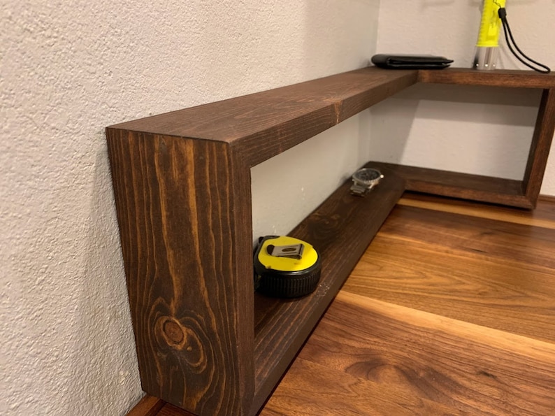 Pu&ograve; includere: Un ripiano d'angolo in legno marrone scuro con due ripiani. Il ripiano superiore ha un portafoglio nero sopra. Il ripiano inferiore ha un metro a nastro giallo e un orologio argentato.