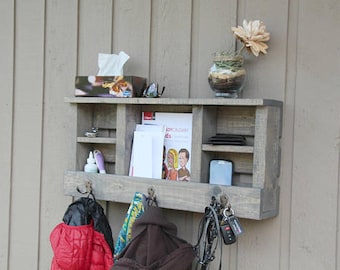 May include: A rustic wooden wall shelf with hooks, three shelves, and a small box on the top shelf. The shelf is painted gray and has a weathered look. There are various items on the shelves, including a box of tissues, a pair of glasses, a book, and a phone. There are also hooks on the bottom shelf with a red jacket, a brown backpack, and a necklace hanging from them.