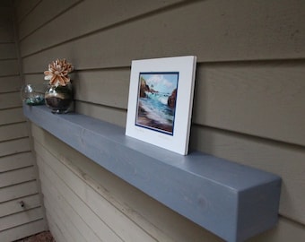 May include: A gray wooden shelf with a seascape painting and two glass vases with decorative elements on top. The shelf is mounted on a light brown wall.