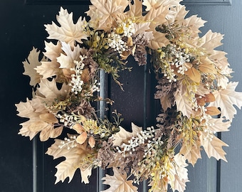 May include: A circular wreath composed of light beige maple leaves, interspersed with small white berries, dark brown dried floral elements, and sprigs of green foliage. The arrangement creates a dense, textured display of autumn-inspired botanical components.