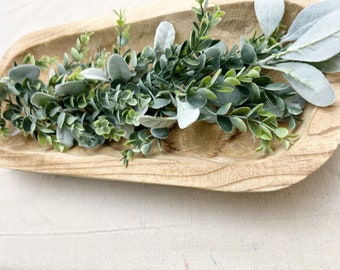 May include: A wooden tray filled with a green and white leafy garland. The garland is made of artificial greenery and has a natural, rustic look.