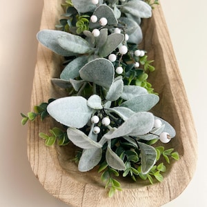 May include: A wooden, oblong decorative bowl filled with faux greenery. The arrangement includes soft, gray-green leaves, small green sprigs, and white berry accents. The bowl has a natural wood grain finish.