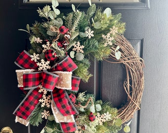 May include: A red and black plaid bow with a burlap accent is attached to a grapevine wreath decorated with greenery and wooden snowflakes.