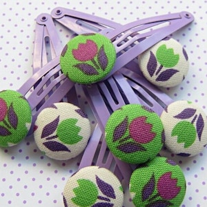 Könnte beinhalten: Ein Satz von acht lilafarbigen Plastikhaarklammern mit stoffbezogenen Knöpfen. Die Knöpfe sind grün mit weißen Mittelpunkten und zeigen ein pinkfarbenes Tulpenmotiv.