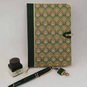 Op de afbeelding: Een groen notitieboek met een gouden en oranje geometrisch patroon op de omslag. Het notitieboek heeft een groene leren band met een gouden gesp en een klein slot met sleutel. Een groene vulpen en een flesje zwarte inkt zijn ook afgebeeld.