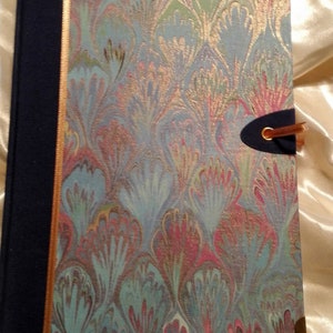 Könnte beinhalten: Ein blaues, grünes und rosafarbenes marmoriertes Tagebuch mit goldener Einfassung und einem blauen Band als Lesezeichen. Das Tagebuch hat einen dekorativen goldenen Verschluss.