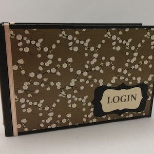 Könnte beinhalten: Ein braunes Buch mit einem weißen Blumenmuster und einem schwarzen Etikett mit der Aufschrift "LOGIN". Das Buch hat ein rosa Band.