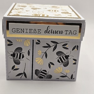 Könnte beinhalten: Eine dekorative Schachtel mit grauem Äußeren, die schwarze und goldene Bienen- und Blumenmuster aufweist. Die Schachtel hat einen goldenen Rand und den Text "GENIEßE deinen TAG" in Schwarz. Die Oberseite der Schachtel hat ein goldenes Bienenmuster.