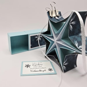Könnte beinhalten: Handgefertigter sternförmiger Schmuck mit einem teal- und weißen Blumenmuster, zusammengehalten mit einer Büroklammer und einem weißen Band. Eine hellblaue Box befindet sich neben dem Schmuck. Eine kleine Karte mit Text ist ebenfalls vorhanden.