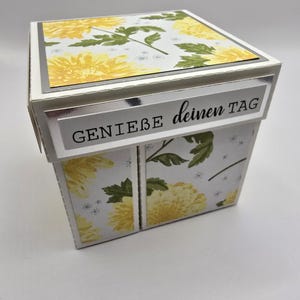Könnte beinhalten: Eine weiße Geschenkbox mit einem floralen Design mit gelben Blumen und grünen Blättern. Die Box hat einen silbernen Rand und den Text "GENIEßE deinen TAG" auf der Vorderseite.