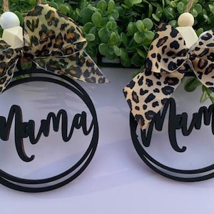 May include: Two black wooden ornaments with the word "Mama" written in a cursive font. Each ornament is decorated with a brown and black leopard print bow.