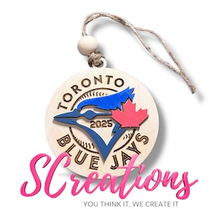 May include: A wooden Toronto Blue Jays ornament with a blue jay logo, a red maple leaf, and the text "2025." The ornament is round with a natural wood finish and a twine hanger. The bottom of the image has the text "SCreations."