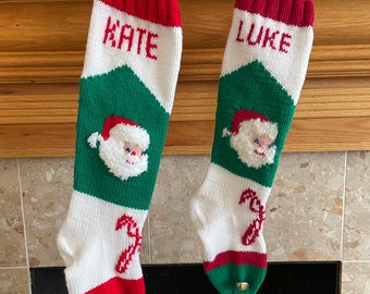 May include: Two red, white, and green knitted Christmas stockings with Santa Claus faces and candy canes. The stockings are personalized with the names "Kate" and "Luke".