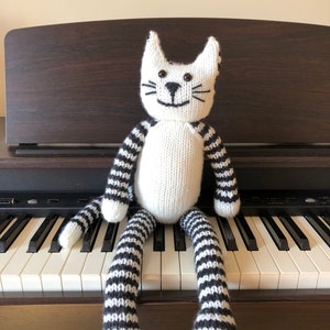 May include: A white knitted cat toy with black stripes sitting on a piano keyboard. The cat has black eyes and a black nose.