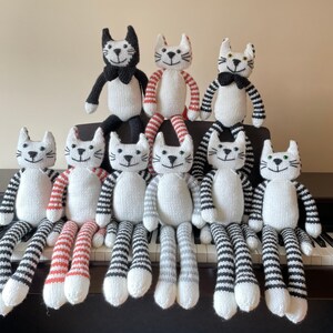 Puede incluir: Un grupo de nueve juguetes de gato tejidos en varios patrones en blanco y negro. Los gatos est&aacute;n todos sentados o de pie sobre una superficie con un teclado en el fondo.