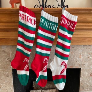 Puede incluir: Tres calcetines navideños personalizados cuelgan de una repisa de madera. Cada calcetín tiene rayas rojas, blancas y verdes con un diseño de bastón de caramelo y un nombre tejido en la parte superior. Los nombres son Patrick, Ayrton y Quinnl.