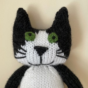 Puede incluir: Un juguete de gato de punto negro y blanco con ojos verdes. El gato tiene la cara y el pecho blancos con orejas, patas y cola negras.