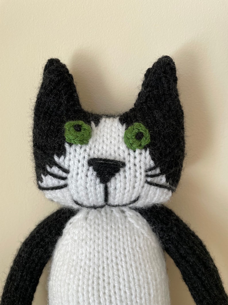 Puede incluir: Un juguete de gato de punto negro y blanco con ojos verdes. El gato tiene la cara y el pecho blancos con orejas, patas y cola negras.