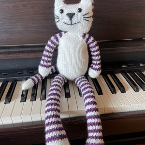 Puede incluir: Un juguete de gato de punto blanco con rayas moradas sentado en un teclado de piano. El gato tiene ojos negros y una nariz negra.