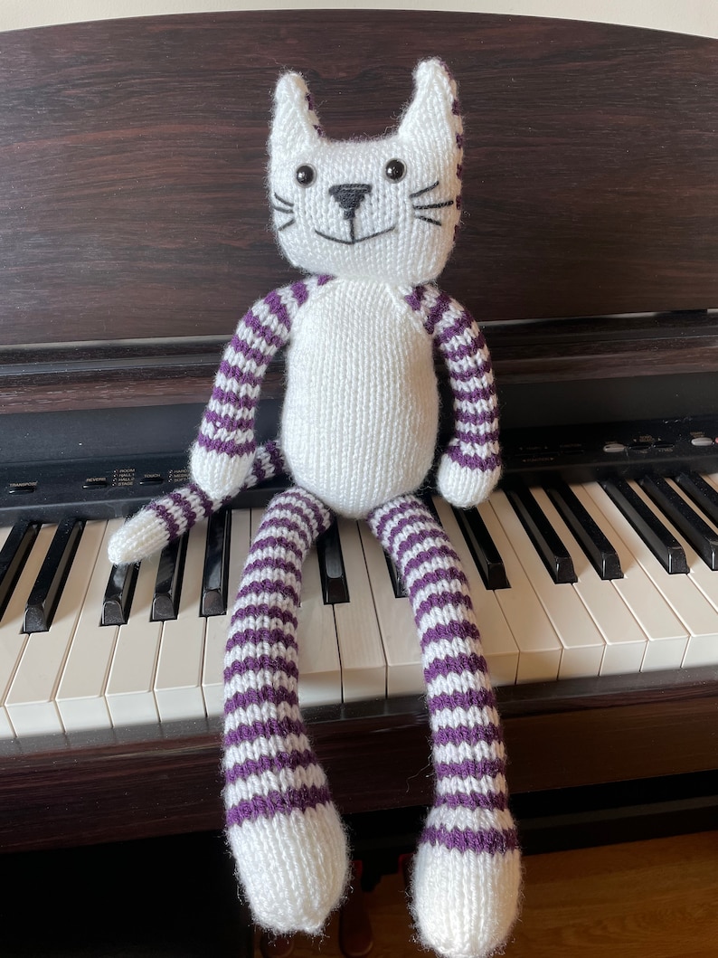 Puede incluir: Un juguete de gato de punto blanco con rayas moradas sentado en un teclado de piano. El gato tiene ojos negros y una nariz negra.