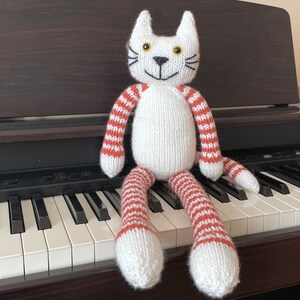 Puede incluir: Un juguete de gato de punto blanco con brazos y piernas a rayas rojas y blancas. El gato est&aacute; sentado en un teclado de piano.