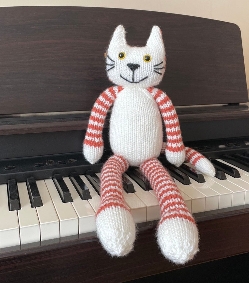 Puede incluir: Un juguete de gato de punto blanco con brazos y piernas a rayas rojas y blancas. El gato est&aacute; sentado en un teclado de piano.
