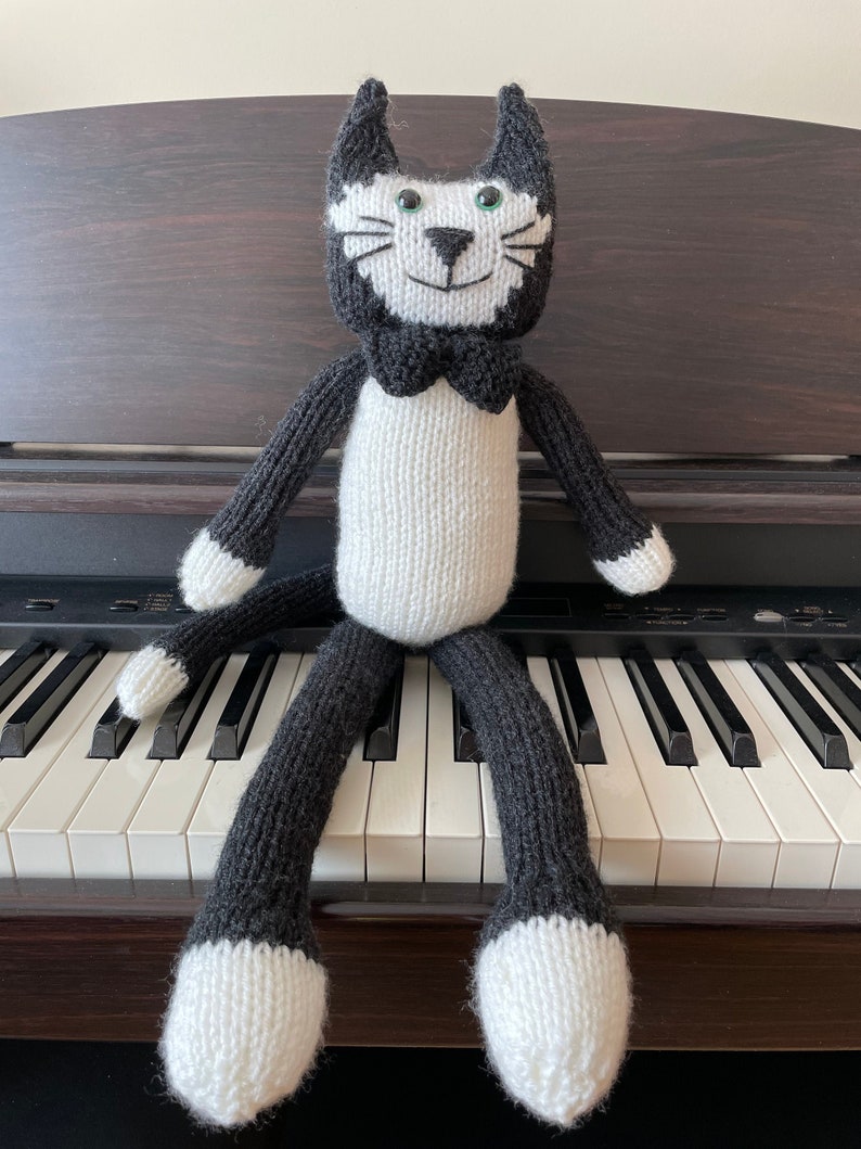 Puede incluir: Un juguete de gato de punto negro y blanco sentado en un teclado de piano. El gato tiene ojos verdes y una pajarita negra.