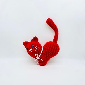 Puede incluir: Un juguete de crochet de gato rojo con un lazo rosa. El gato está sentado con la cola enroscada alrededor de su cuerpo.
