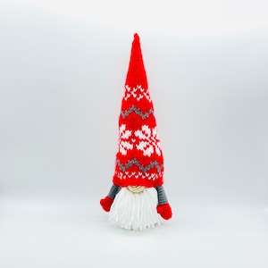 Puede incluir: Un gnomo de punto rojo con un patrón de copo de nieve blanco y gris en el gorro. El gnomo tiene una barba blanca y guantes rojos.