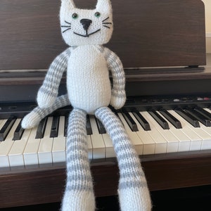 Puede incluir: Un juguete de gato de punto blanco con rayas grises en los brazos y las piernas. El gato est&aacute; sentado en un teclado blanco y negro.