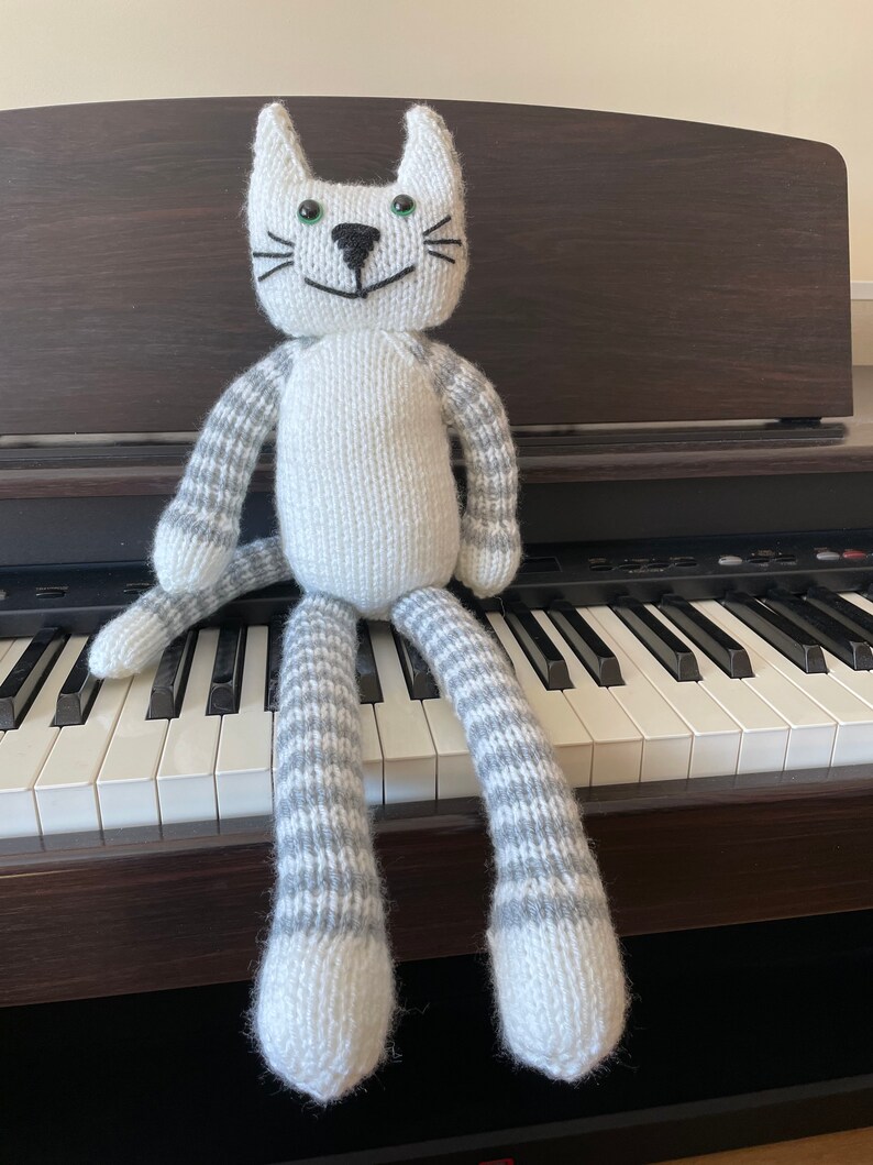 Puede incluir: Un juguete de gato de punto blanco con rayas grises en los brazos y las piernas. El gato est&aacute; sentado en un teclado blanco y negro.