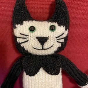 Puede incluir: Un juguete de gato de punto blanco y negro con ojos verdes. El gato tiene un collar negro y est&aacute; sentado sobre una superficie roja.