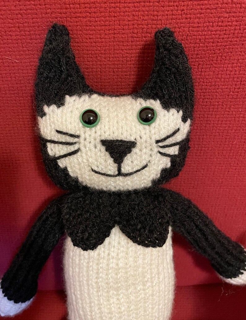 Puede incluir: Un juguete de gato de punto blanco y negro con ojos verdes. El gato tiene un collar negro y est&aacute; sentado sobre una superficie roja.