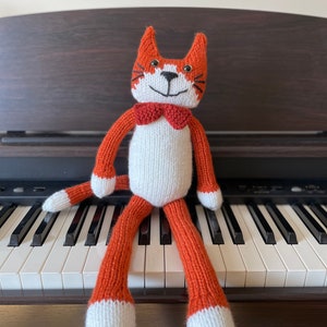 Puede incluir: Un juguete de gato de punto naranja y blanco con una pajarita negra sentado en un teclado de piano blanco y negro.
