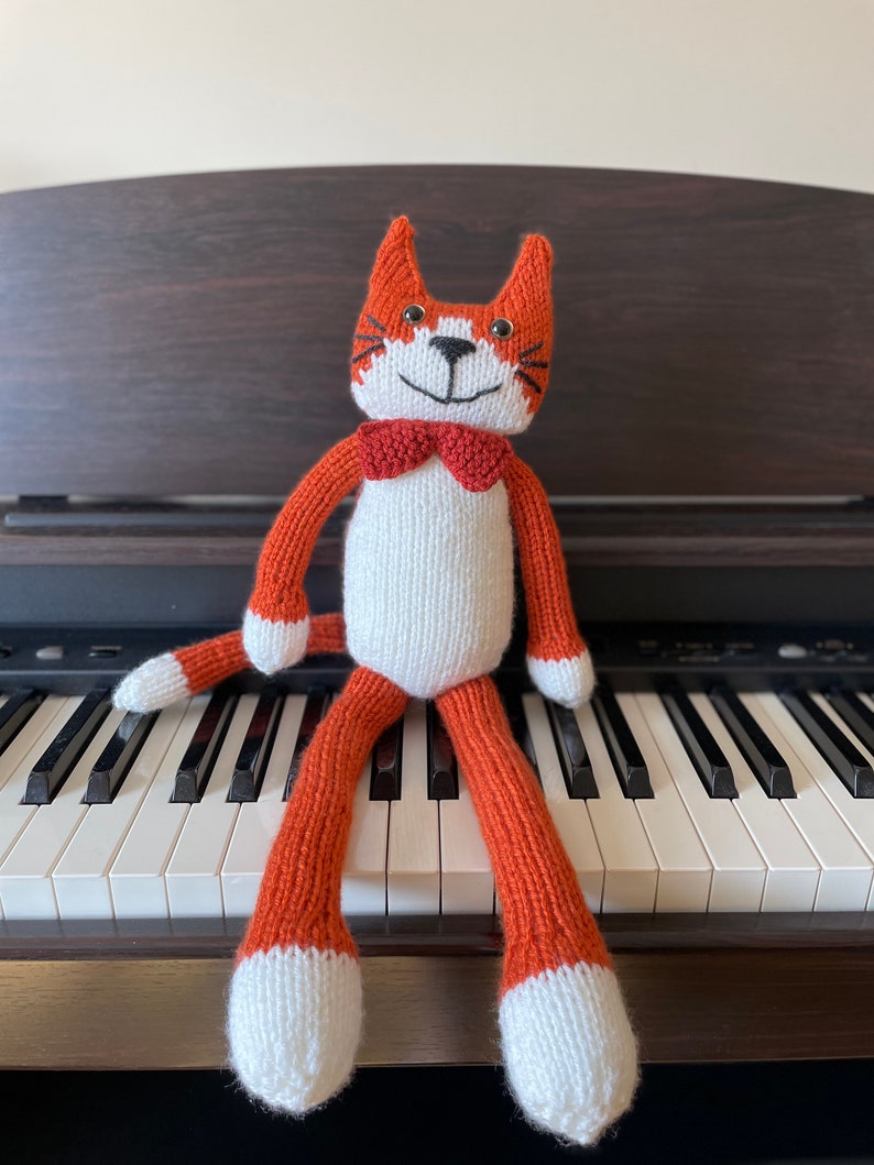 Puede incluir: Un juguete de gato de punto naranja y blanco con una pajarita negra sentado en un teclado de piano blanco y negro.