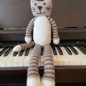 Puede incluir: Un juguete de gato de punto gris y blanco con ojos verdes y nariz negra sentado en un teclado de piano.