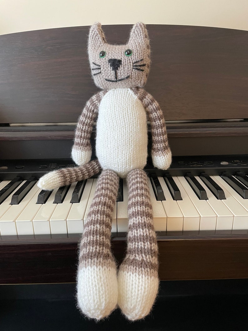 Puede incluir: Un juguete de gato de punto gris y blanco con ojos verdes y nariz negra sentado en un teclado de piano.