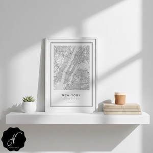 May include: A framed black and white map of New York City, with the city name at the bottom. The map is displayed on a white shelf, alongside a small succulent in a white pot, a stack of books, and a cork-topped cup.