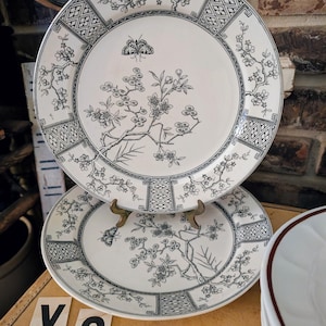 May include: Two white ceramic plates with a black floral and butterfly design. The plates have a decorative border and are displayed on a wooden surface. The plates are part of a dinnerware set.