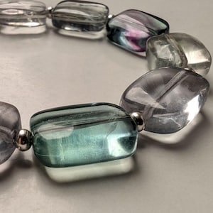 Könnte beinhalten: Ein Armband aus rechteckigen, durchscheinenden Perlen in klaren, hellblauen und violetten Farbtönen, die durch kleine, polierte silberfarbene Metallzwischenperlen getrennt sind. Die Perlen haben eine glatte Oberfläche und sind kreisförmig angeordnet.