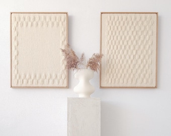 Puede incluir: Dos tapices de pared blancos enmarcados con un diseño texturizado, colgados en una pared blanca sobre un pedestal blanco con un jarrón de cerámica blanco y hierba de pampa seca.
