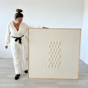 May include: A large, square, off-white textured artwork in a light wooden frame. The artwork features a vertical arrangement of raised, rectangular shapes. The piece is displayed in a bright, white room.