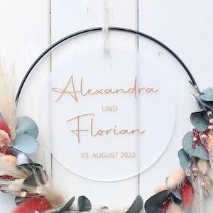 Könnte beinhalten: Ein weißes rundes Acrylschild mit einem schwarzen Metallreifen und einem Blumenkranz. Das Schild trägt die Aufschrift "Alexandra und Florian 03. August 2022".