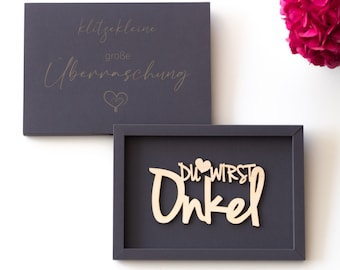 Könnte beinhalten: Eine graue Box mit goldener Beschriftung, die "klitzekleine große Überraschung" und ein kleines Herz lautet. Eine zweite graue Box mit einem Holzrahmen enthält ein Holzschild mit der Aufschrift "Du wirst Onkel" mit einem kleinen Herz.